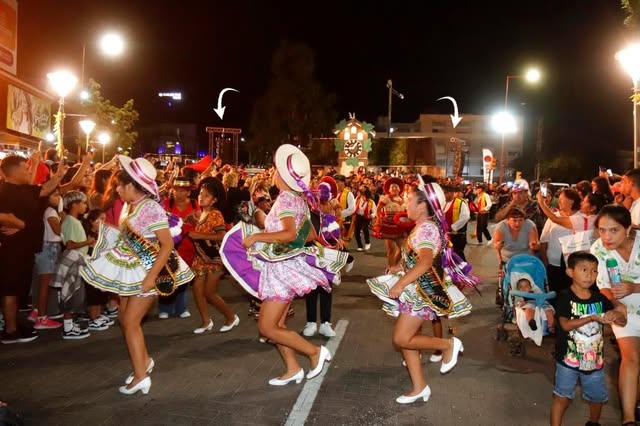 Evento al aire libre con público numeroso y comparsa bailando en la calle iluminada.