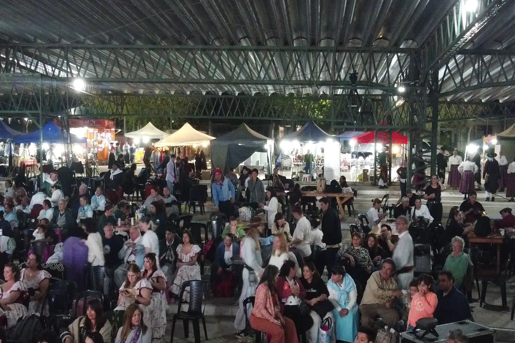 Tribuna colmada de público en un evento masivo, iluminada por las luces del predio de noche.