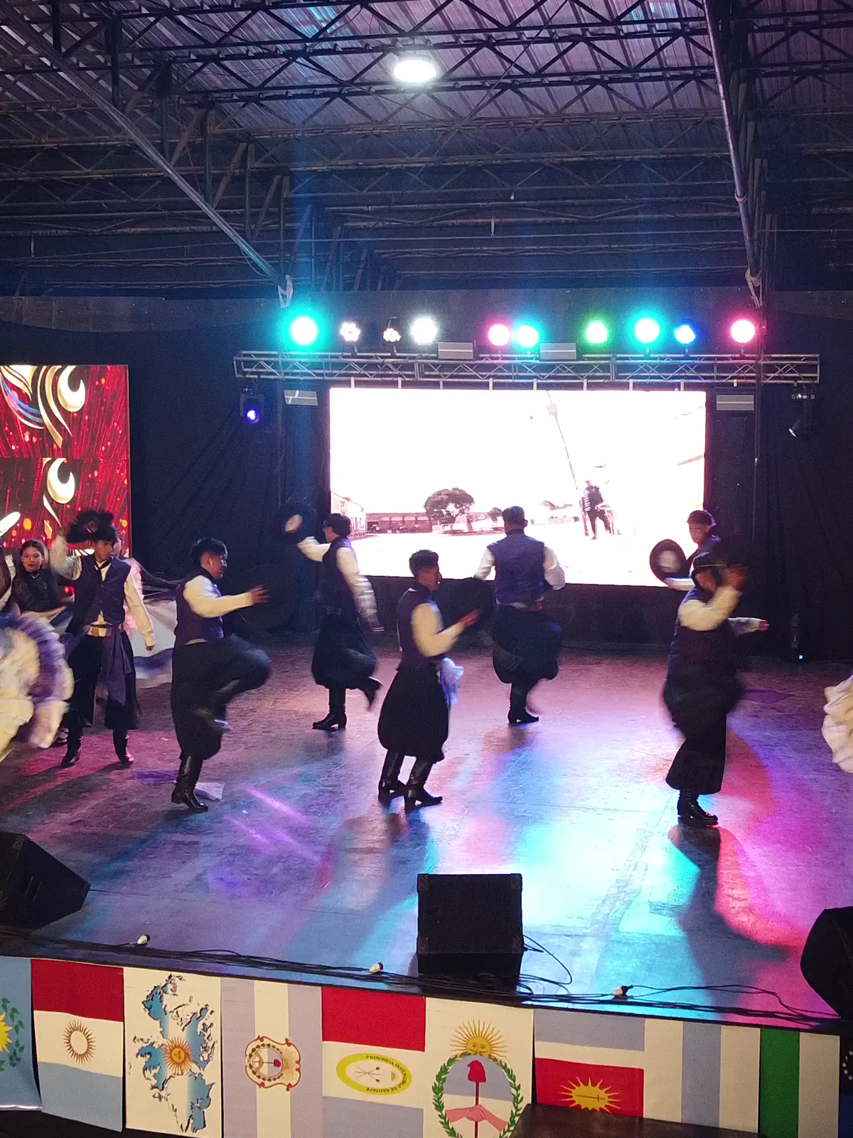 Grupo de baile en pleno salto sobre el escenario, con pantalla LED y truss de luces de colores encendido.